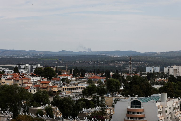 A view of the skyline from Israel looking into Lebanon. Some buildings can be seen in the foreground, but in the distance, smoke rises in a pillar against hilly terrain.