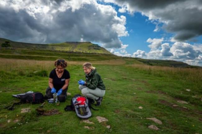 Two women sit on the ground with sampling equipment in a green landscape