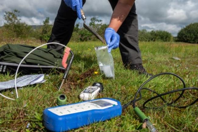 A meter lies on the ground in the foreground while a person wearing gloves takes a sample behind