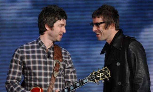 Liam and Noel Gallagher together in 2008, photographed against a blue background; Noel wears a checked shirt and holds a guitar, while Liam, who is smiling, wears sunglasses and a black leather jacket